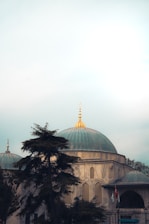 A mosque with a large, ornate dome topped with a gold finial, surrounded by trees. The dome and structure are made of stone with arched windows featuring lattice designs. Flags are visible near the entrance.