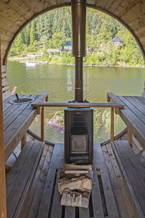 A rustic sauna interior with wooden benches on either side and a central stove. The space is enclosed with a large, round window offering a scenic view of a lake and lush greenery. Logs of wood are placed in front of the stove, ready for use. The view outside includes a riverbank with thick, green trees and a nearby structure across the water.