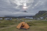 A camping site in a mountainous area with an orange tent in the foreground and several caravans and vehicles parked along grassy fields. Low-lying clouds drift across the mountain tops, partially obscuring them, while the sun peeks through the cloudy sky, casting a soft light over the landscape.