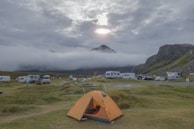 A camping site in a mountainous area with an orange tent in the foreground and several caravans and vehicles parked along grassy fields. Low-lying clouds drift across the mountain tops, partially obscuring them, while the sun peeks through the cloudy sky, casting a soft light over the landscape.