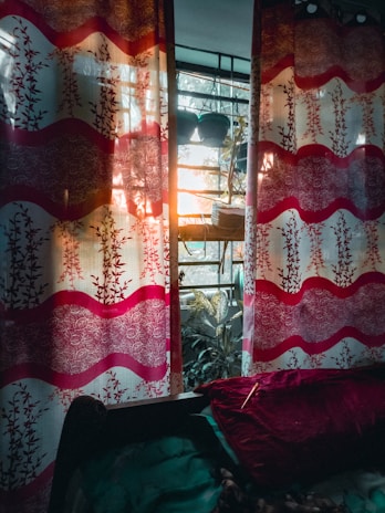 A vibrant, patterned curtain with red and white floral designs hangs on a window. The sunlight filters through the fabric, creating a warm glow. Behind the curtain, green potted plants rest on a windowsill, and a garden can be seen outside. A red velvet pillow and a dark-colored bedspread are visible in the lower part of the image.