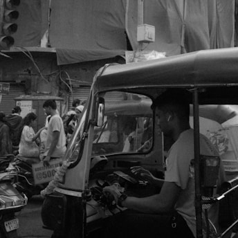 A busy urban street scene with a focus on a man sitting inside an auto-rickshaw, interacting with a device. In the background, several pedestrians are walking, and some are engaged in conversation. The area appears to be densely packed, likely in a market or commercial district. Overhead, tarps and wires hang from buildings, contributing to a cluttered atmosphere.