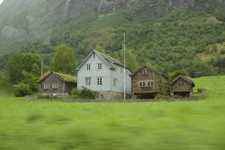 A group of rustic wooden houses with various shades of brown and a central blue wooden house are surrounded by lush greenery. The houses are positioned closely together, with a picket fence enclosing them. The background consists of dense forested hills and mountains, covered with green foliage.