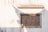 A decorative wooden window with intricate carvings set into a white, slightly weathered wall. The carvings depict floral and geometric patterns. Shadows cast by nearby structures create interesting angles and shapes on the wall.