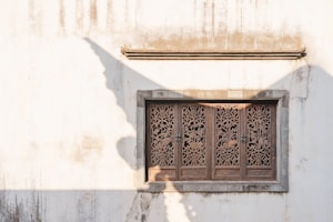 A decorative wooden window with intricate carvings set into a white, slightly weathered wall. The carvings depict floral and geometric patterns. Shadows cast by nearby structures create interesting angles and shapes on the wall.