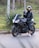 A person wearing a black leather jacket and a reflective helmet is sitting on a sleek black motorcycle parked on the side of a road. The person is making a peace sign with their hand. The surroundings are green and lush, with trees and vegetation in the background.