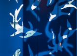 A modern abstract artwork featuring white organic and leaf-like shapes against a vibrant blue background. The design is reminiscent of nature, with light appearing to pass through the shapes, creating dynamic shadows.