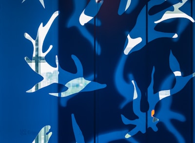 A modern abstract artwork featuring white organic and leaf-like shapes against a vibrant blue background. The design is reminiscent of nature, with light appearing to pass through the shapes, creating dynamic shadows.