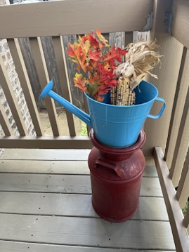 A blue watering can is filled with autumn-themed decorations, including artificial orange and yellow leaves as well as dried corn cobs. This watering can is placed on top of a rustic red milk can, all situated on a wooden deck that is surrounded by a railing and part of a fence visible in the background.