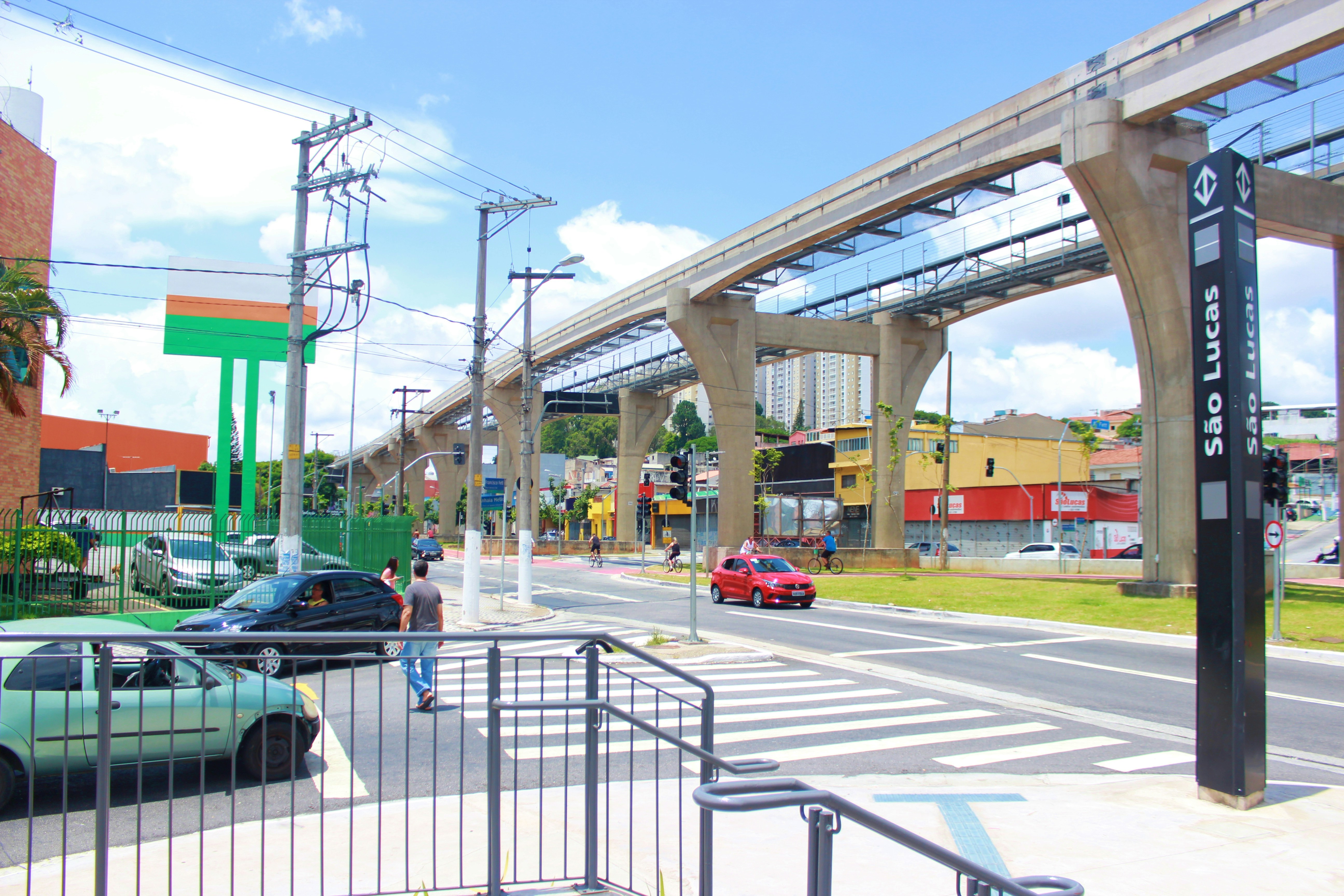 Via elevada do monotrilho sobre uma avenida, com pilares de concreto, carros, faixa de pedestres e a placa da estação