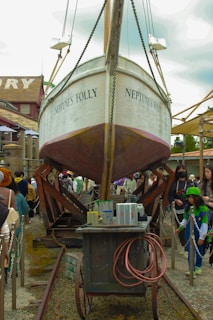 A large boat named 'Neptune's Folly' is suspended in the air with ropes and supports. It is centered among a bustling crowd of people within an outdoor setting. There is a cart with various cans of paint, a coiled hose, and other equipment beneath the boat. The sky is overcast, creating a diffused lighting effect.