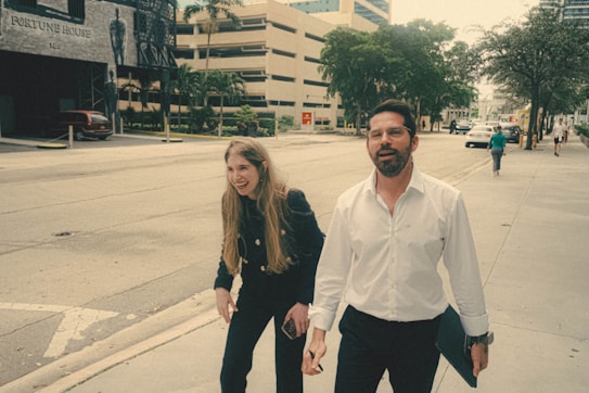 Two people are walking on a city sidewalk, enjoying a conversation. The person on the left has long hair and is dressed in dark clothing, while the person on the right is wearing a white shirt and holding a folder. The background shows a street with trees, a building labeled 'Fortune House,' and a few parked cars.