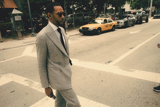A man in a gray suit and sunglasses walks across a street with a yellow taxi in the background. He holds a phone in his hand. The scene appears to be urban, with several parked cars and trees lining the street.