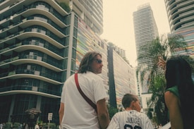 A group of people stands in an urban environment surrounded by tall, modern buildings with distinctive colorful patterns. One person appears to be pointing towards something, possibly explaining or showing something to others. The environment is lush with palm trees amidst the skyscrapers, and the overall scene is vibrant and dynamic.