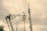 A utility worker is elevated in a bucket truck, working on overhead electrical wires and equipment attached to a tall utility pole. The background displays overcast skies with a greyish hue. A construction crane and other utility poles are visible in the distance.