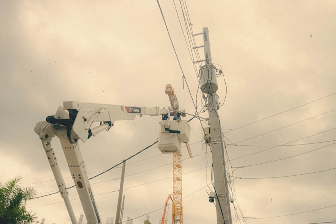 A utility worker is elevated in a bucket truck, working on overhead electrical wires and equipment attached to a tall utility pole. The background displays overcast skies with a greyish hue. A construction crane and other utility poles are visible in the distance.