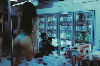 A dimly lit convenience store with a person in a blue outfit standing in the foreground. Behind them, another person is seated near refrigerated shelves filled with various drinks. The scene appears casual and candid, with the surrounding area crowded with beverage bottles.