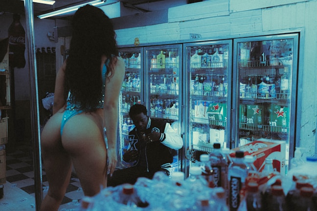 A dimly lit convenience store with a person in a blue outfit standing in the foreground. Behind them, another person is seated near refrigerated shelves filled with various drinks. The scene appears casual and candid, with the surrounding area crowded with beverage bottles.