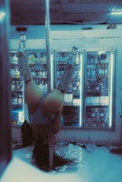 A person is performing an inverted dance move on a pole in a dimly lit room with fluorescent lighting. The background features a refrigerated display full of various bottles, suggesting a setting like a convenience store or liquor shop. The floor is scattered with what appears to be paper currency.