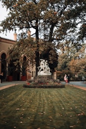 A stone sculpture is positioned in the center of a well-maintained garden surrounded by lush greenery and blooming flowers. Tall trees with dense foliage form a backdrop, set against a historic building with arched windows and decorative brickwork. People are passing by, adding a sense of life and movement to the peaceful scene.