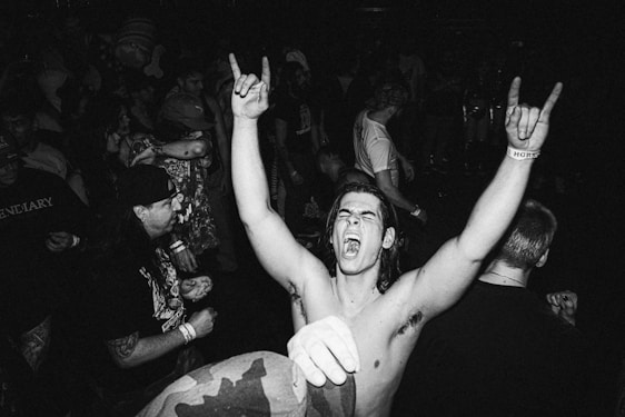 A man in the foreground is raising his arms with clenched fists, displaying a rock hand gesture and appearing to shout in excitement. A crowd of people surrounds him, engaging in what seems to be a live music event or concert. The image is in black and white, adding a classic or vintage feel.