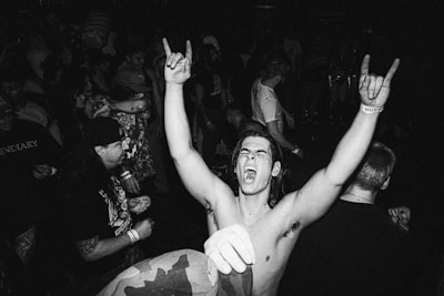 A man in the foreground is raising his arms with clenched fists, displaying a rock hand gesture and appearing to shout in excitement. A crowd of people surrounds him, engaging in what seems to be a live music event or concert. The image is in black and white, adding a classic or vintage feel.