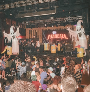 A lively concert scene with a band performing on stage. The stage is adorned with inflatable ghost figures and barrels with a Halloween theme. The crowd is animated, enjoying the music with some audience members dressed in festival attire. There is vibrant lighting and an energetic atmosphere.