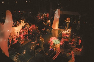 A concert is taking place with a band performing on a stage in front of a large crowd. The stage is dimly lit with a warm, yellowish lighting. Large inflatable ghost figures are positioned around the stage, adding to the overall atmosphere. The band members appear energetic, engaging with the audience, and playing various instruments including drums and guitars. The crowd is packed and seems to be enjoying the performance.