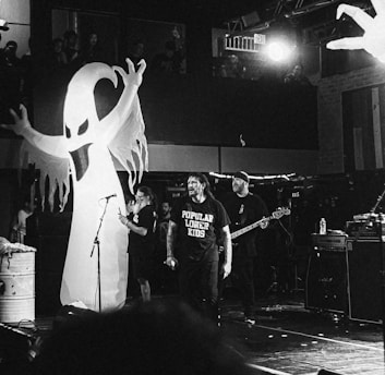 A band is performing on stage with a large inflatable ghost figure in the background. Several musicians are present, including one with a guitar and another with a microphone. The setting appears to be a concert venue with some audience members visible in an upper gallery.