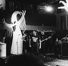 A band is performing on stage with a large inflatable ghost figure in the background. Several musicians are present, including one with a guitar and another with a microphone. The setting appears to be a concert venue with some audience members visible in an upper gallery.