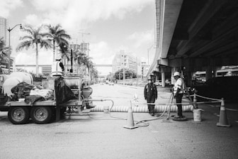 Workers are engaged in roadwork under an overpass with construction equipment and traffic cones. Palm trees and city buildings are visible in the background, along with a partially cloudy sky. One worker stands on a trailer filled with equipment, while another worker operates a tool on the ground. The third worker is standing near the middle, observing the work.