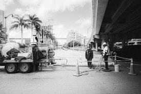 Workers are engaged in roadwork under an overpass with construction equipment and traffic cones. Palm trees and city buildings are visible in the background, along with a partially cloudy sky. One worker stands on a trailer filled with equipment, while another worker operates a tool on the ground. The third worker is standing near the middle, observing the work.