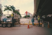 A street under an overpass features a construction site with several workers wearing reflective vests and hard hats. Traffic cones and safety barriers are arranged around the area. A trailer with equipment and materials is parked nearby, and palm trees line the side of the street. In the background, a building under construction and cranes are visible.
