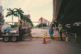 A street under an overpass features a construction site with several workers wearing reflective vests and hard hats. Traffic cones and safety barriers are arranged around the area. A trailer with equipment and materials is parked nearby, and palm trees line the side of the street. In the background, a building under construction and cranes are visible.