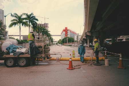 A street under an overpass features a construction site with several workers wearing reflective vests and hard hats. Traffic cones and safety barriers are arranged around the area. A trailer with equipment and materials is parked nearby, and palm trees line the side of the street. In the background, a building under construction and cranes are visible.