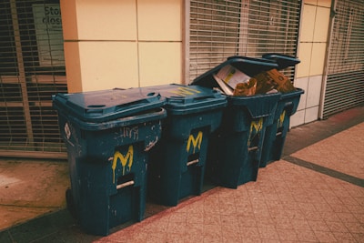 Three blue recycling bins are positioned on a sidewalk against a building with closed metal shutters. The bins display a yellow 'M' logo and the one on the far right is overfilled with cardboard boxes.