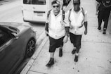 Two men wearing safety vests and hats walk along a sidewalk beside parked vehicles. The image is in black and white, with one man wearing sunglasses and both carrying backpacks.