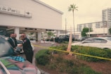A police officer stands next to a car outside an automotive service and parts center, interacting with two men in formal attire. Yellow caution tape surrounds a small landscaped area, and several cars are parked in the background. A building with large glass windows is visible in the distance.