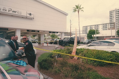 A police officer stands next to a car outside an automotive service and parts center, interacting with two men in formal attire. Yellow caution tape surrounds a small landscaped area, and several cars are parked in the background. A building with large glass windows is visible in the distance.