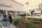 A police officer stands next to a car outside an automotive service and parts center, interacting with two men in formal attire. Yellow caution tape surrounds a small landscaped area, and several cars are parked in the background. A building with large glass windows is visible in the distance.