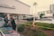 A police officer stands next to a car outside an automotive service and parts center, interacting with two men in formal attire. Yellow caution tape surrounds a small landscaped area, and several cars are parked in the background. A building with large glass windows is visible in the distance.