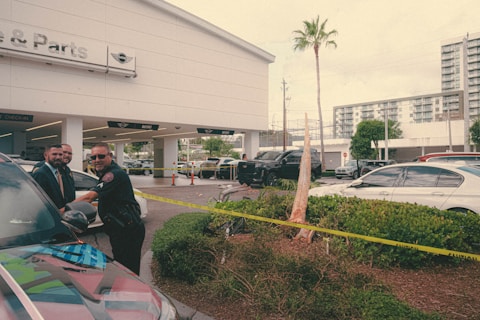 A police officer stands next to a car outside an automotive service and parts center, interacting with two men in formal attire. Yellow caution tape surrounds a small landscaped area, and several cars are parked in the background. A building with large glass windows is visible in the distance.