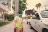 A construction worker wearing a high-visibility vest, face covering, and protective gloves stands near a white utility pickup truck with traffic cones in the background. The urban setting includes tall buildings, palm trees, and a traffic sign displaying information.