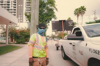 A construction worker wearing a high-visibility vest, face covering, and protective gloves stands near a white utility pickup truck with traffic cones in the background. The urban setting includes tall buildings, palm trees, and a traffic sign displaying information.