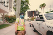 A construction worker wearing a high-visibility vest, face covering, and protective gloves stands near a white utility pickup truck with traffic cones in the background. The urban setting includes tall buildings, palm trees, and a traffic sign displaying information.