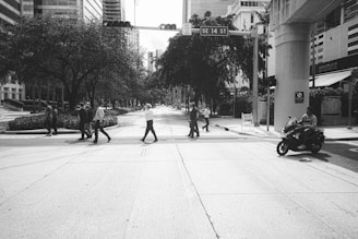 A group of people crosses a city street at an intersection lined with tall buildings. Trees provide shade along the sidewalks, and there is a person riding a scooter. The area appears to be business-oriented, with people dressed in business attire.