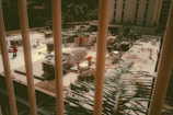A construction site seen through vertical bars features workers in orange safety vests carrying out various tasks. The site is organized with materials and equipment scattered around. In the background, buildings and trees are visible, adding an urban setting.