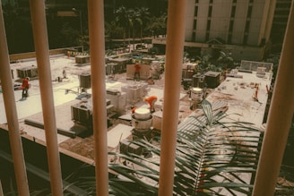 A construction site seen through vertical bars features workers in orange safety vests carrying out various tasks. The site is organized with materials and equipment scattered around. In the background, buildings and trees are visible, adding an urban setting.