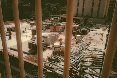 A construction site seen through vertical bars features workers in orange safety vests carrying out various tasks. The site is organized with materials and equipment scattered around. In the background, buildings and trees are visible, adding an urban setting.