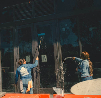 Three individuals engaged in cleaning large glass doors. One person is using a broom or mop to scrub the glass, another is spraying water, and the third person is wearing a hard hat and a safety vest. There are construction materials in the foreground.